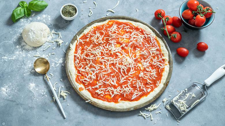 Pizza on blue surface with dough, cheese, tomatoes, herbs, and kitchen tools