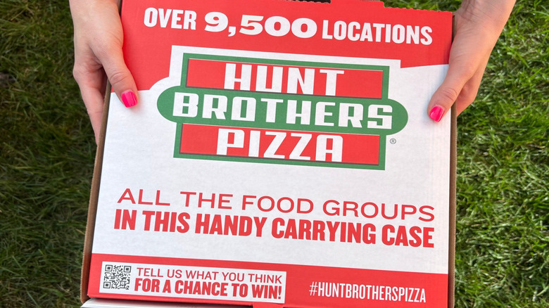 Hands holding a Hunt Brothers Pizza box over a lawn