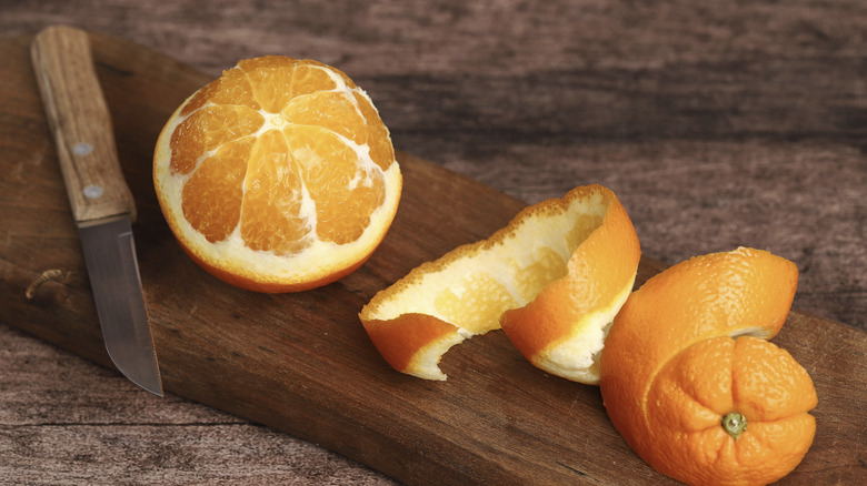 A half-peeled orange sitting on a cutting board next to a knife and the discarded peel.