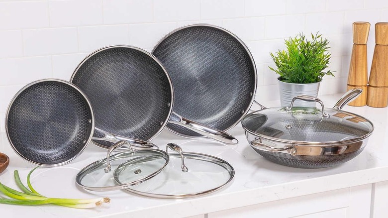 Four HexClad pans and lids sitting on a white counter next to green onions and a potted herb