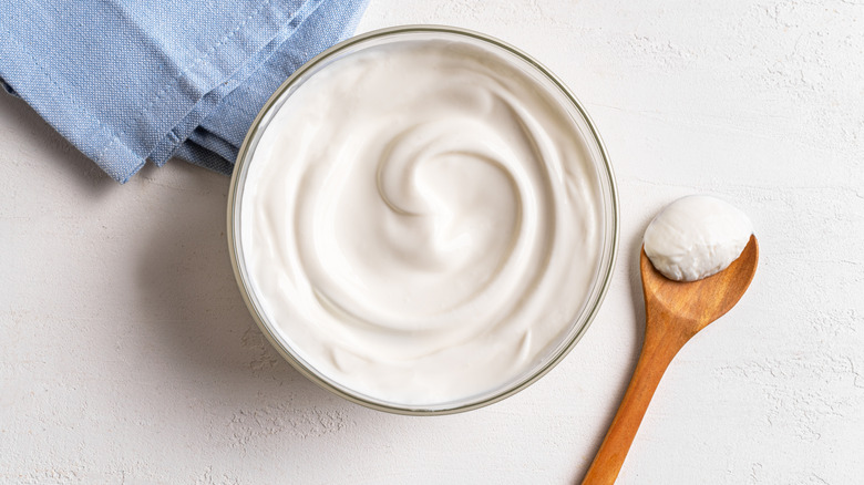 Glass bowl filled with plain yogurt on white background next to blue linen napkin and wooden spoon