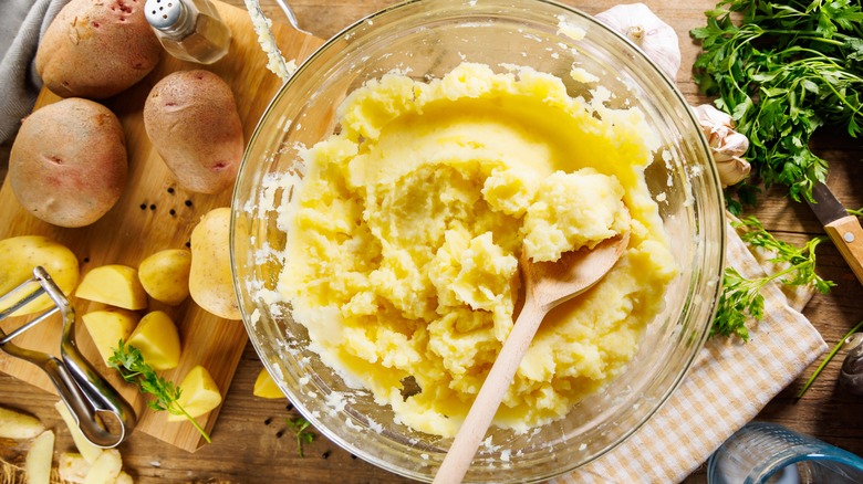Mashed potatoes in a bowl next to cut and peeled potatoes