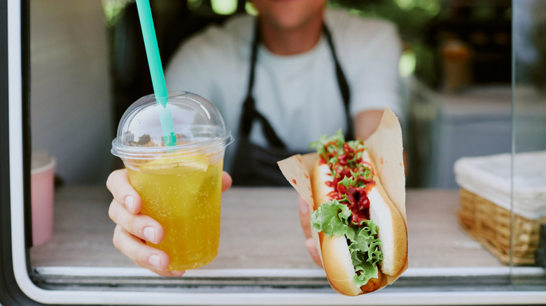 Close up of a lemonade and a hot dog in hands of unrecognizable chef at fast food establishment