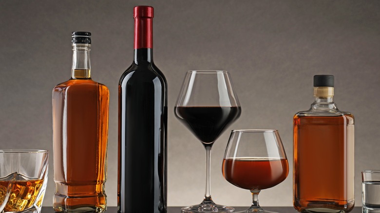 Bottles of whiskey and wine are lined up by a tumbler of whiskey, a stemmed glass of red wine, and a snifter of whiskey