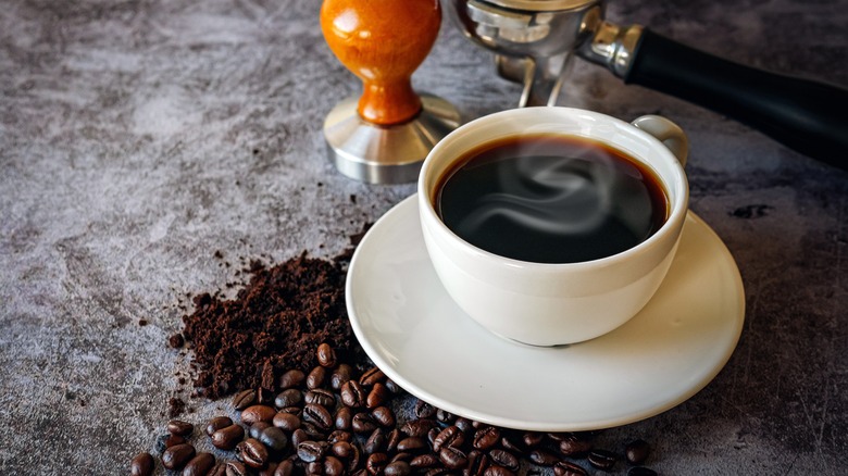 A steaming cup of coffee surrounded by coffee grounds, coffee beans, and coffee brewing utensils.